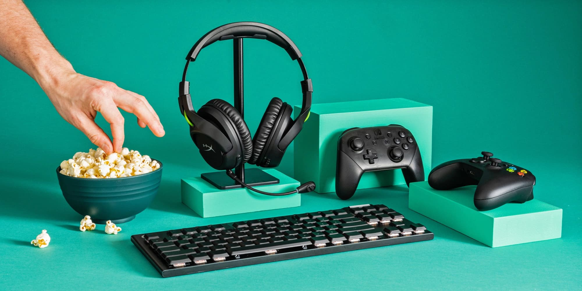 Gaming accessories including a headset, controllers, and mechanical keyboard arranged on a teal background.